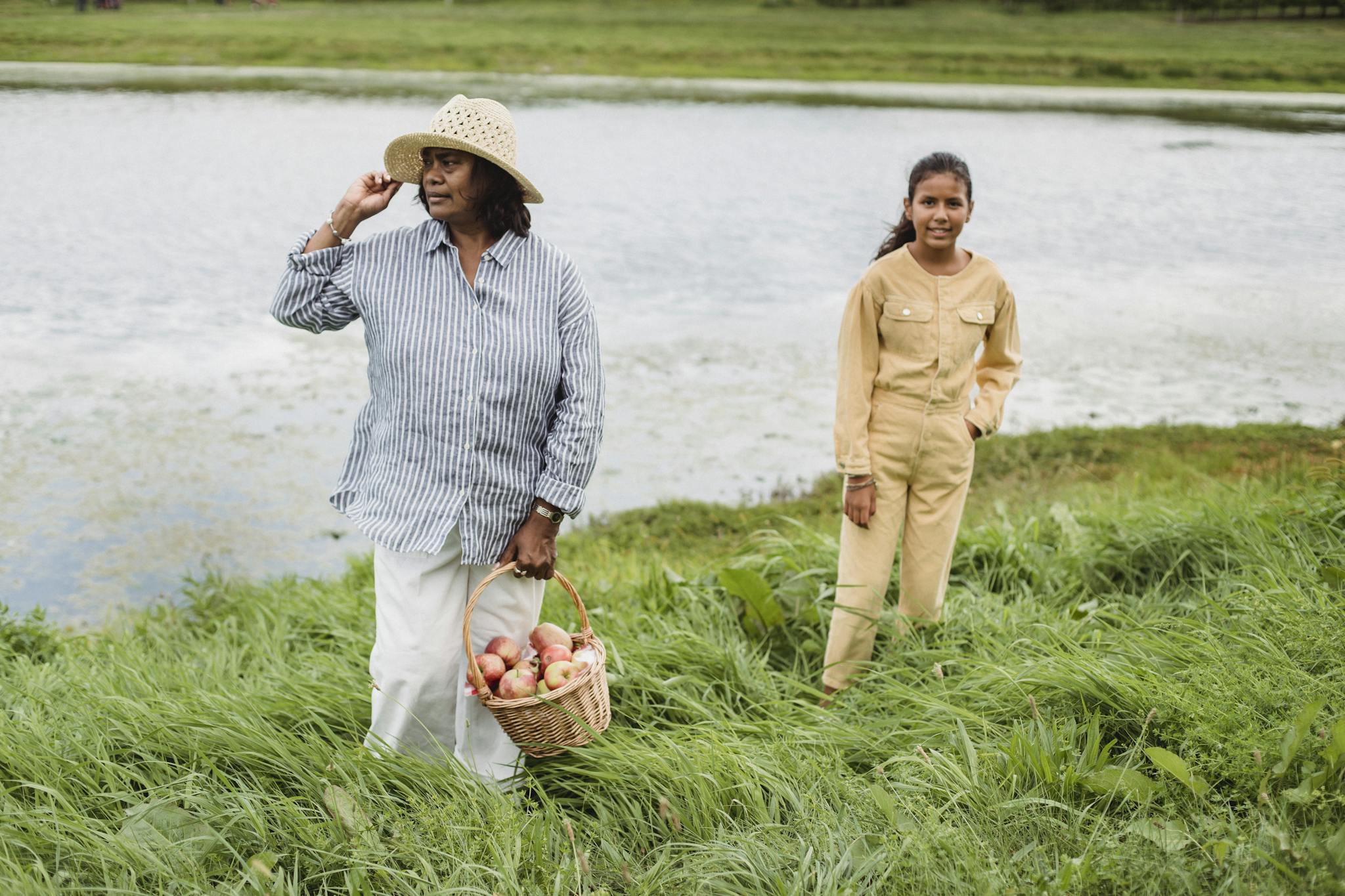 A woman and a young girl enjoy harvesting apples outdoors near a serene pond.